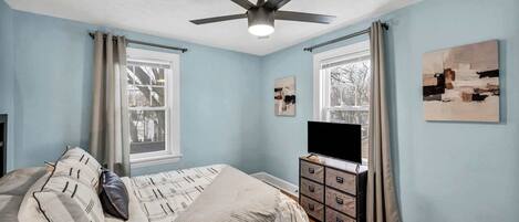 A bedroom with a bed, a ceiling fan, a TV, and artwork on the walls.