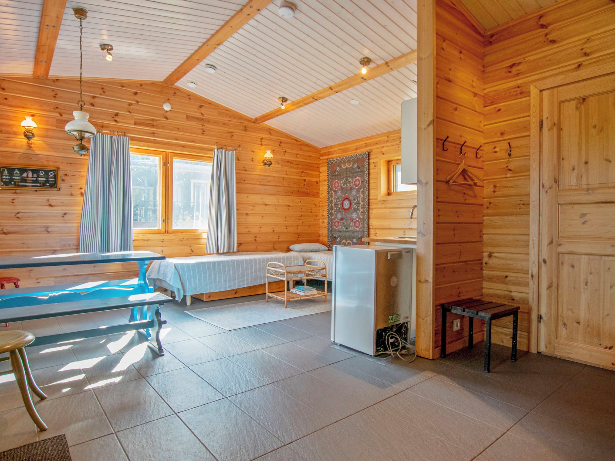 A cozy cabin interior with a bed, a dining table, a small refrigerator, and a window with curtains.