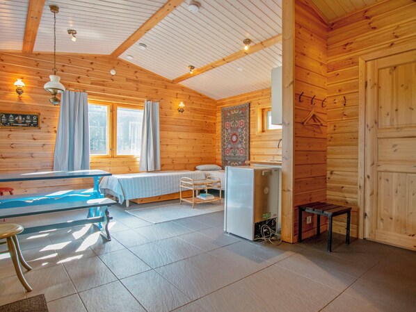 A cozy cabin interior with a bed, a dining table, a small refrigerator, and a window with curtains.