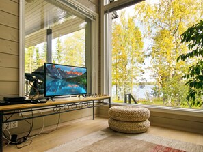 A room with a large window, a television displaying a scenic landscape, a chessboard, and a woven ottoman.