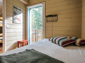 A wooden cabin room with a bed, a small table, a poster on the wall, and a window with a view of trees.