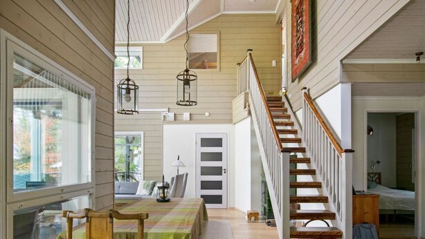 Una habitación amplia con una escalera de madera, un comedor con mesa y sillas, y vistas al exterior a través de amplios ventanales.