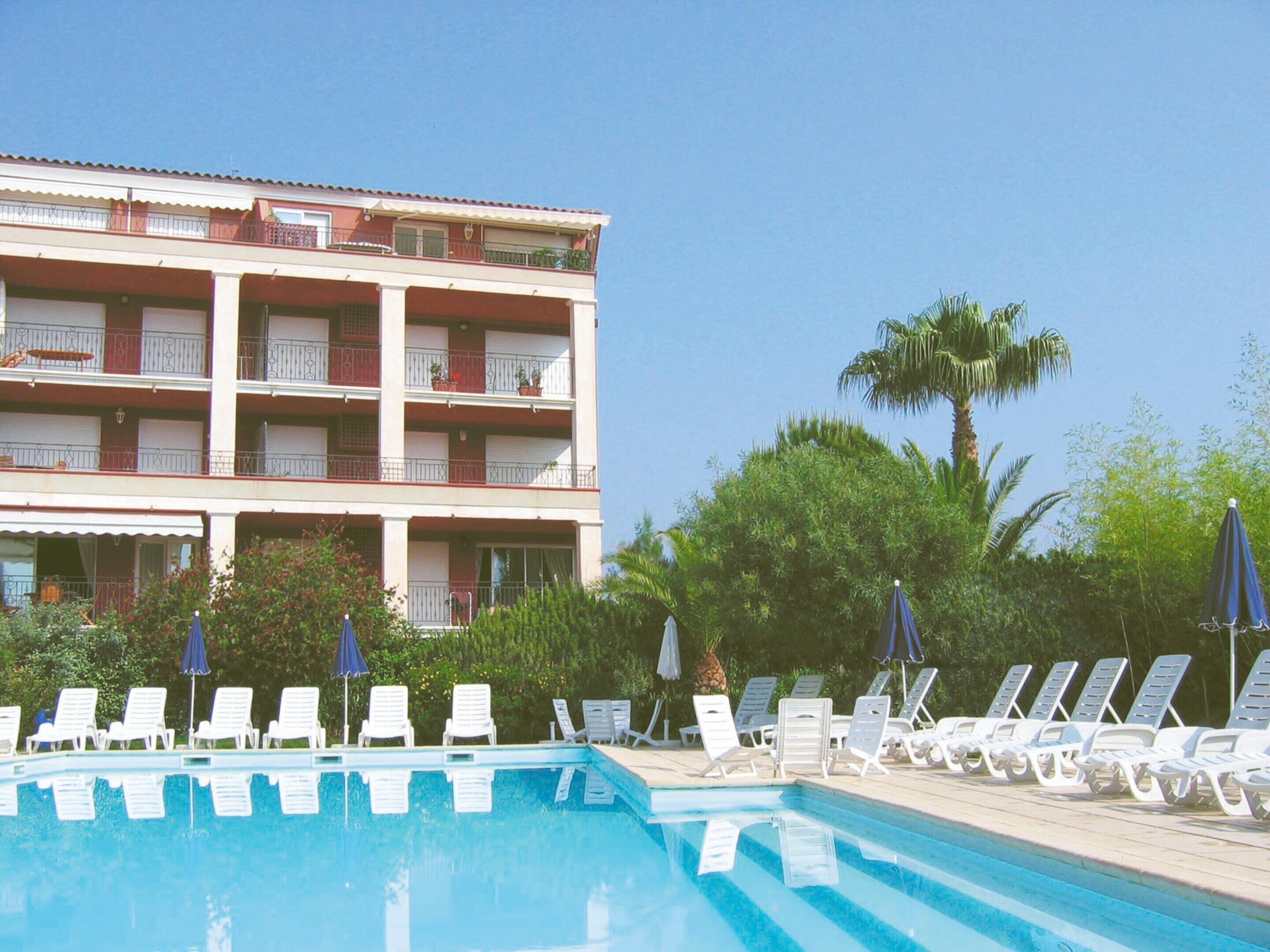 Un espace piscine avec des chaises longues et des parasols, un immeuble à plusieurs étages doté de balcons et une végétation tropicale.