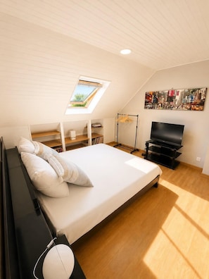 Un dormitorio con cama, televisor, repisa, perchero y tragaluz.