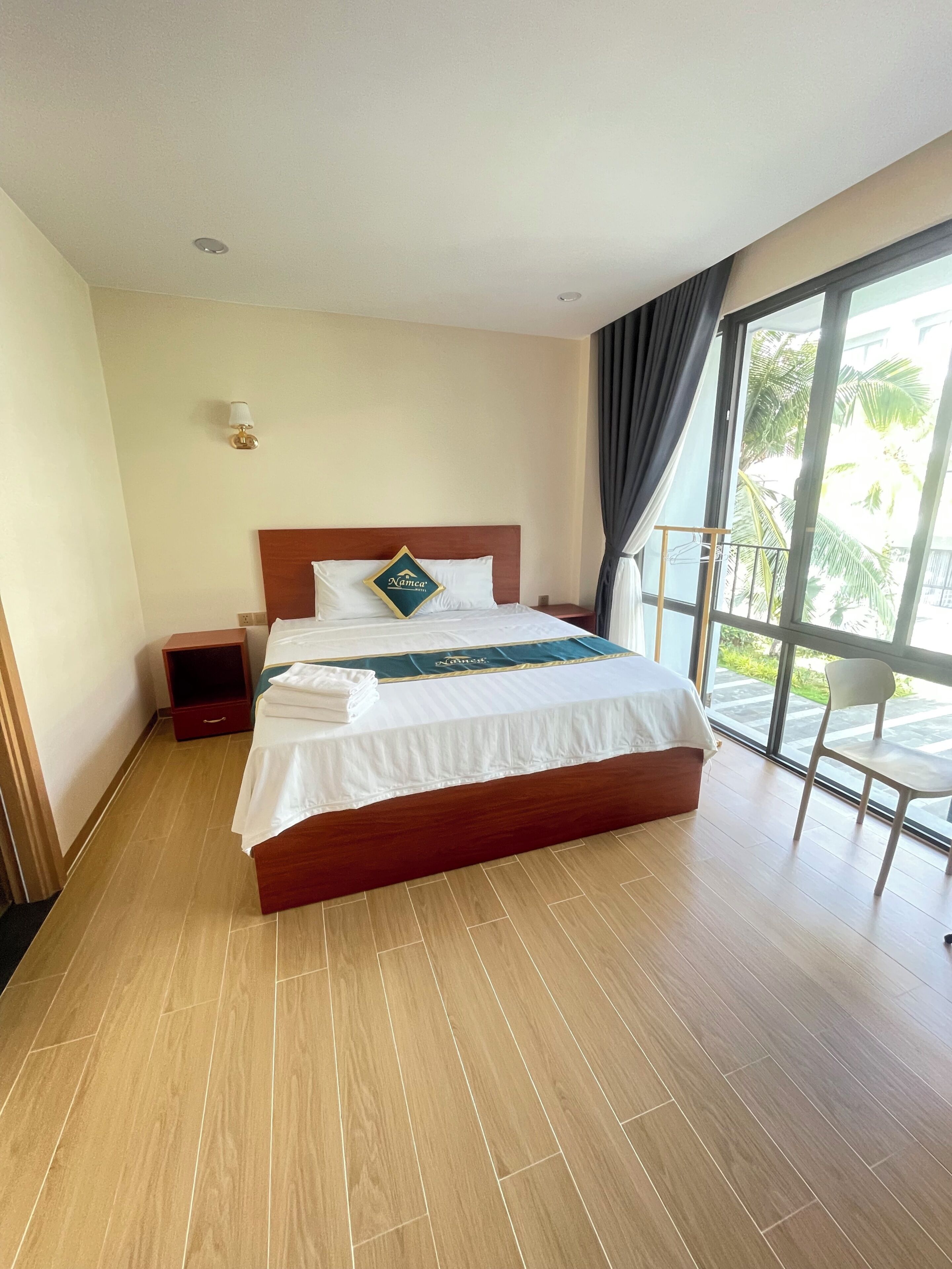 Habitación de hotel con una cama de madera, una mesita de noche, una lámpara y vistas a palmeras a través de un amplio ventanal.