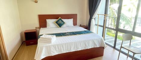 Habitación de hotel con una cama de madera, una mesita de noche, una lámpara y vistas a palmeras a través de un amplio ventanal.