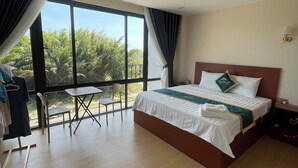 A hotel room with a large bed, a small table, a chair, and a view of palm trees through large windows. - Namca Hotel  (Phu Quoc)