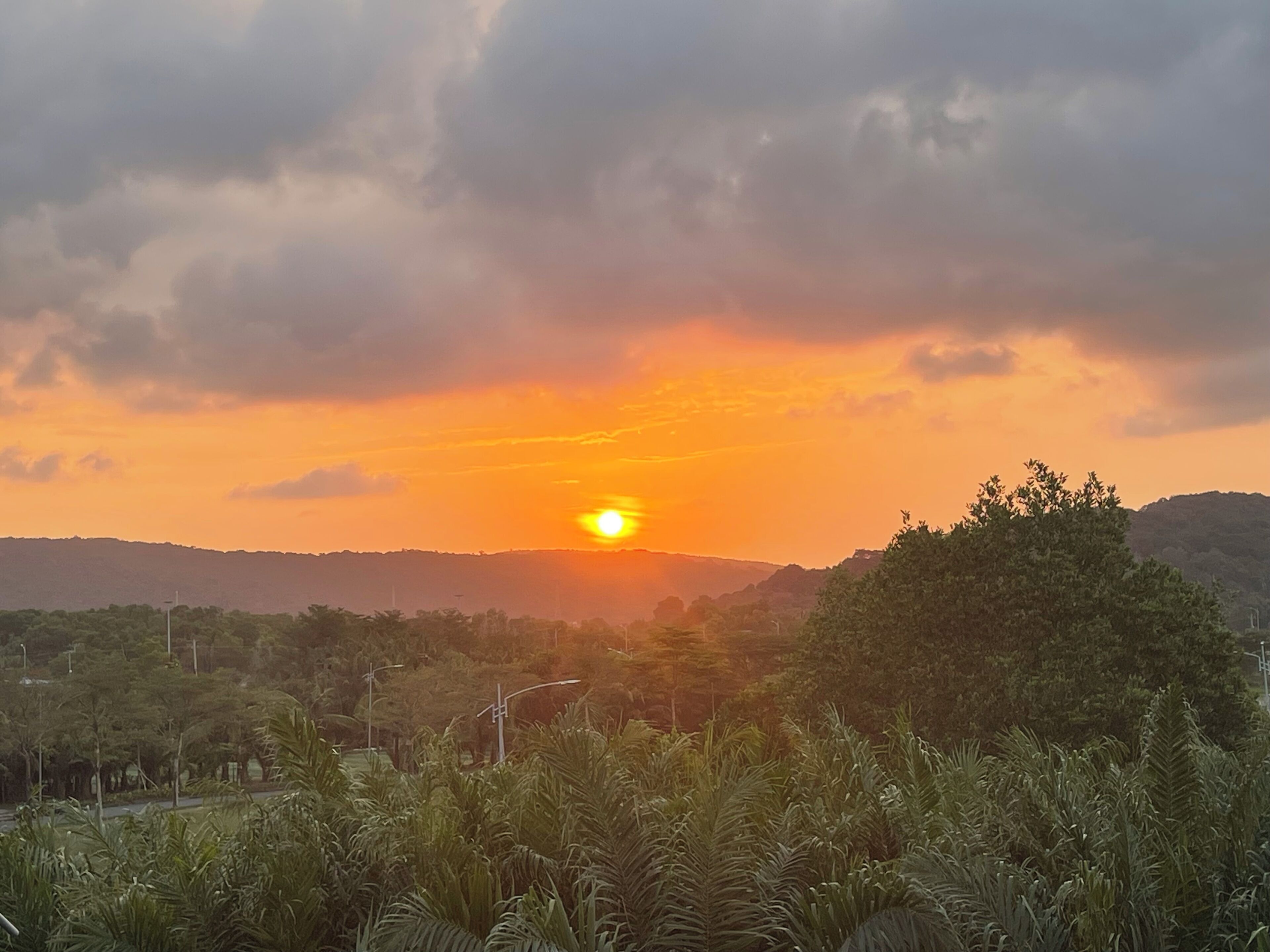 A sunset over a hilly landscape with trees and streetlights.