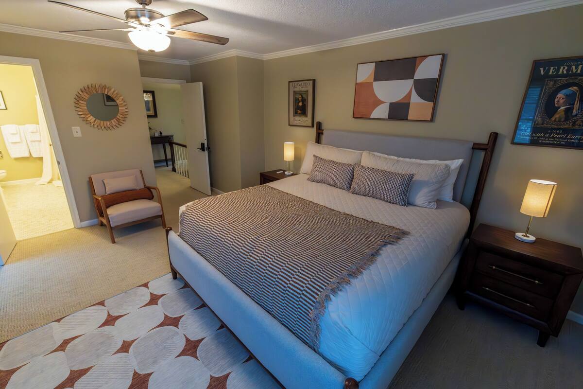 A bedroom with a large bed, a chair, a bedside table, a lamp, a ceiling fan, and a framed picture on the wall.