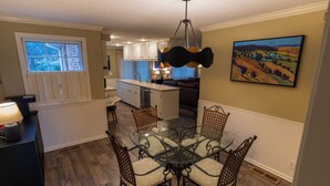 A dining area with a glass table and metal chairs, a kitchen in the background, and a painting on the wall. - The Cunningham (Salina)