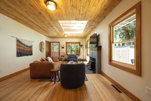 A cozy living room with wooden floors, a fireplace, and large windows.