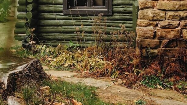 A log cabin with a stone chimney, a window with a grid pattern, and a wheelbarrow in the foreground.