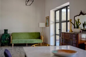A room with a green sofa, a wooden chair, a table with a bowl, a lamp, a vase, and a window with blinds.