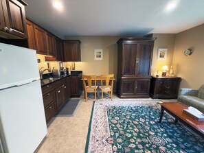 A kitchen with wooden cabinets, a white refrigerator, a dining table with chairs, a large dark wooden cabinet, and a patterned rug.