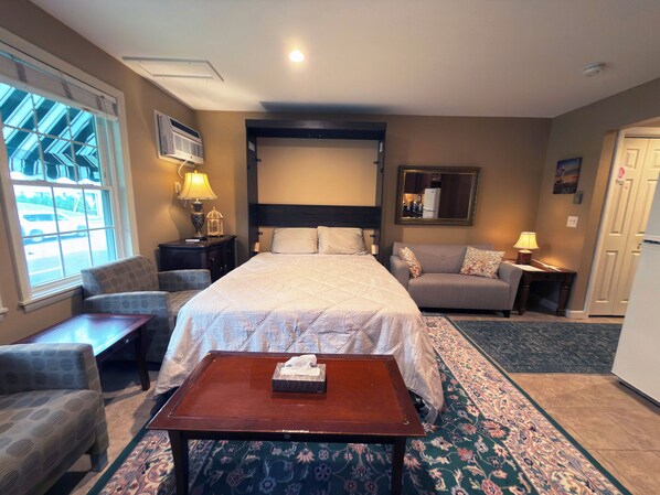 A hotel room with a bed, a sofa, a small table, a lamp, a mirror, and a window with a view of the outside.