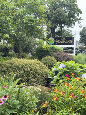 Ein Schild des Hotels 'The Seasons', umgeben von üppigem Grün und blühenden Pflanzen.