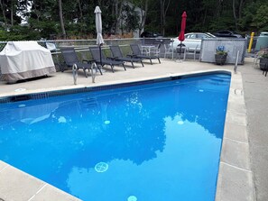 Ein Schwimmbecken mit Liegestühlen und Sonnenschirmen, umgeben von einem Zaun und Bäumen.