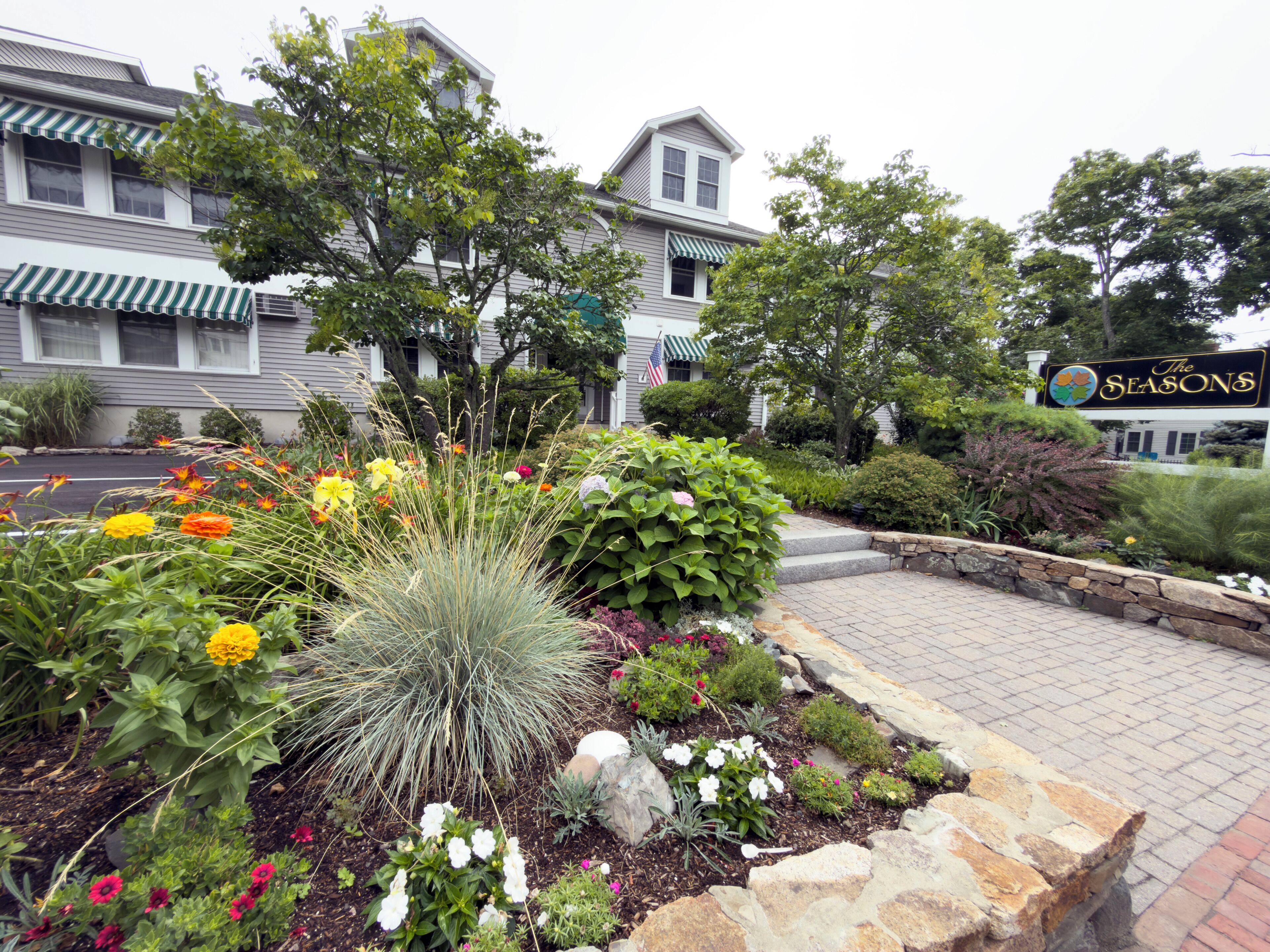 A landscaped garden with a variety of plants and flowers, a stone pathway, and a building with a sign that reads 'The Seasons.'