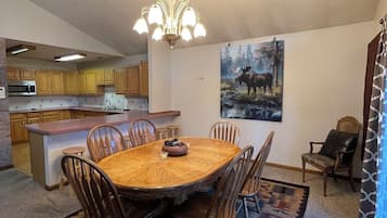 Un coin repas avec une table ronde, des chaises, un lustre, une cuisine en arrière-plan et un tableau au mur.