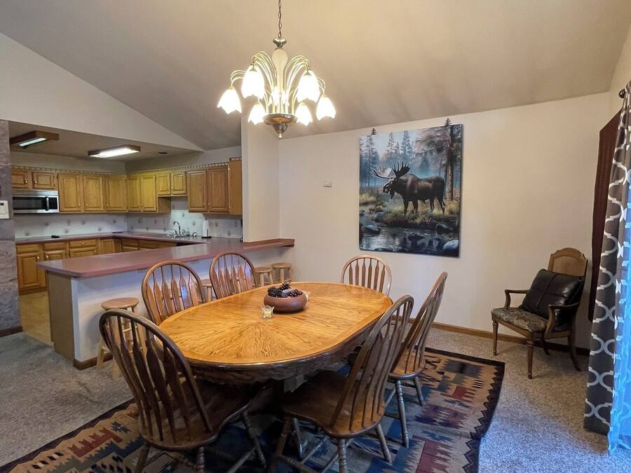 Un coin repas avec une table ronde, des chaises, un lustre, une cuisine en arrière-plan et un tableau au mur.
