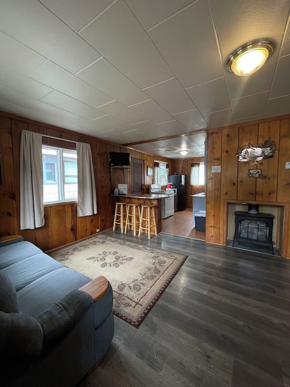 A cabin interior with a sofa, wooden floors, a kitchen area with a stove, and a fireplace.