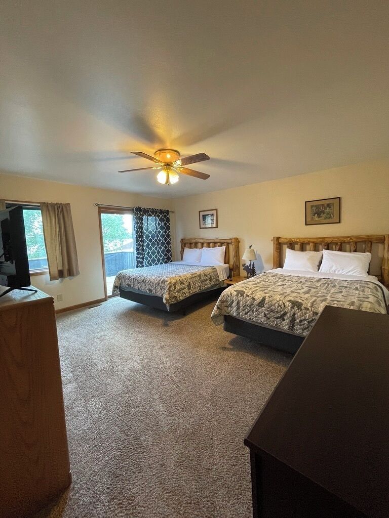 A hotel room with two beds, a ceiling fan, a television, and a window with curtains.