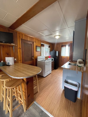 A compact kitchen with a stove, microwave, and sink. There is a wooden table with stools, and a trash bin is visible.