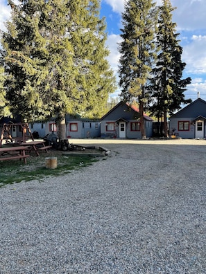 A gravel parking area with two cabins, each with a red-framed window and a door, surrounded by tall evergreen trees.