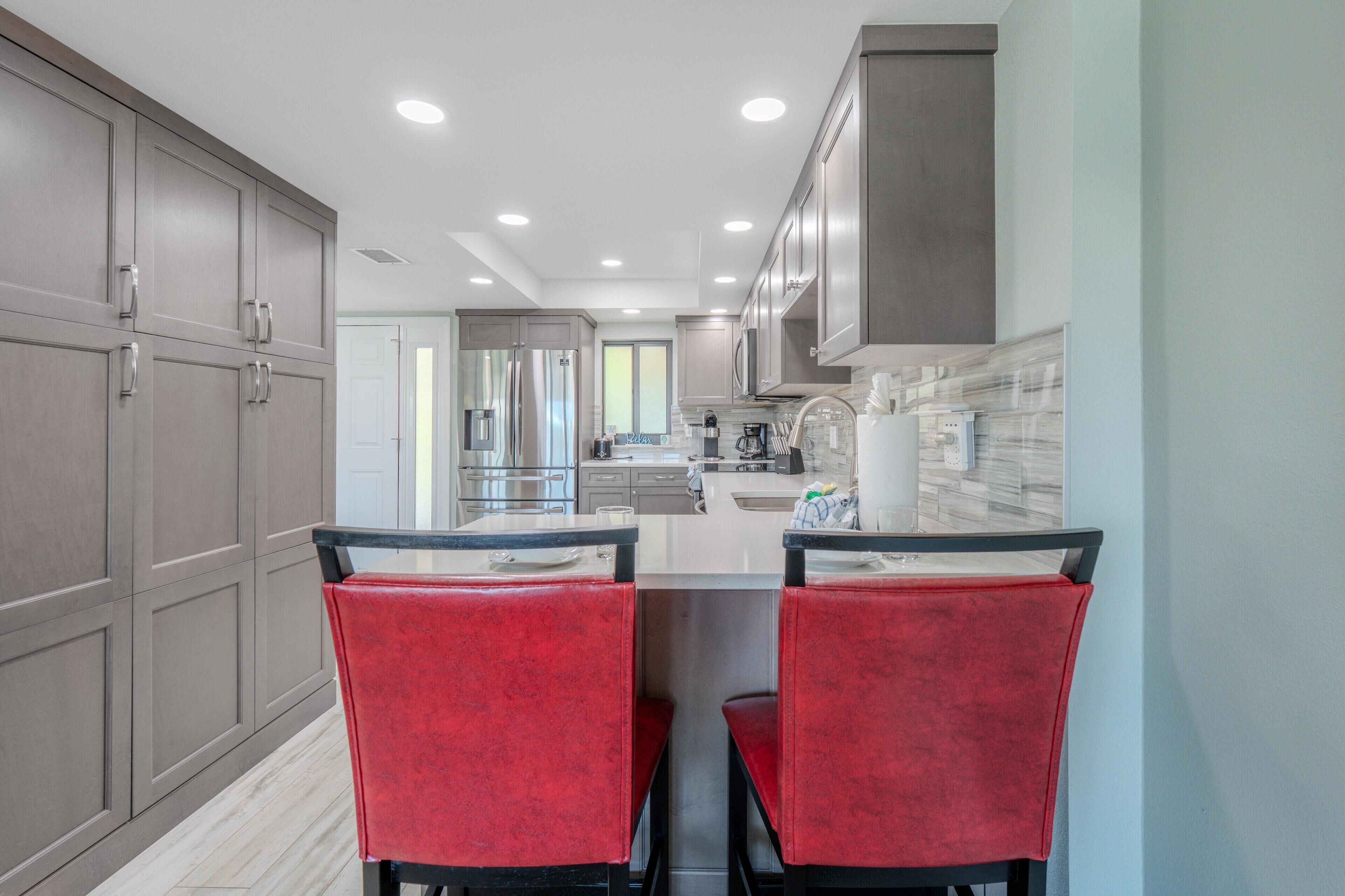 A modern kitchen with two red chairs, stainless steel appliances, and grey cabinetry.