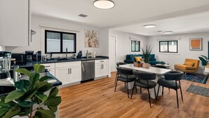 Een moderne keuken met witte kasten, een eethoek met een tafel en stoelen, en een woonkamer met een bank en een gele fauteuil.