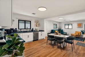 Una cocina moderna con armarios blancos, un comedor con mesa y sillas, y una sala de estar con sofá y un sillón amarillo.