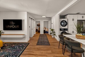 Una cocina moderna con armarios blancos, un comedor con mesa y sillas, un televisor montado en la pared y un pasillo que conduce a un dormitorio.