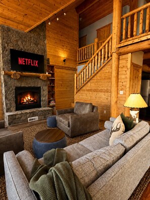 A cozy log cabin interior with a stone fireplace, a Netflix sign, a staircase, and a comfortable seating area. - Luxury Off-Grid Mountain Cabin on 4 Acres, Views, Serenity and Wellness (Jefferson)
