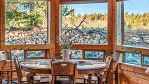 A wooden interior with a round dining table, chairs, and large windows offering a view of a rocky landscape and trees. - Luxury Off-Grid Mountain Cabin on 4 Acres, Views, Serenity and Wellness (Jefferson)