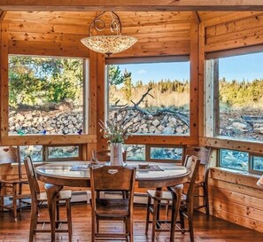 A wooden interior with a round dining table, chairs, and large windows offering a view of a rocky landscape and trees. - Luxury Off-Grid Mountain Cabin on 4 Acres, Views, Serenity and Wellness (Jefferson)