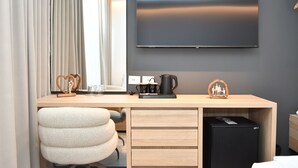 Habitación de hotel con televisión de pantalla plana, escritorio de madera con cafetera, refrigerador pequeño y una silla cómoda.