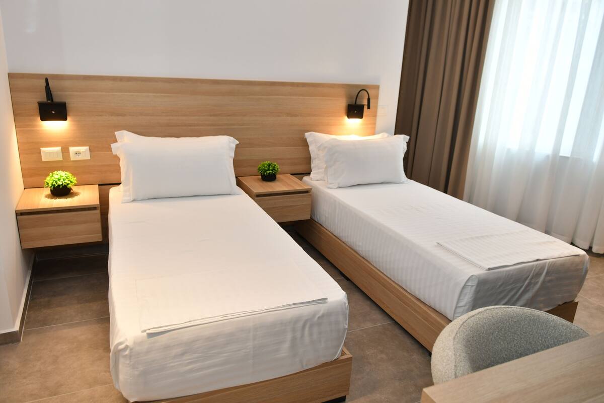 Una habitación de hotel con dos camas individuales, cabeceros de madera, mesitas de noche con lámparas, una pequeña planta en maceta y una ventana con cortinas.