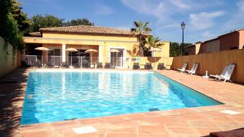 A rectangular swimming pool with clear blue water, surrounded by lounge chairs and umbrellas, and a building with a tiled roof in the background.