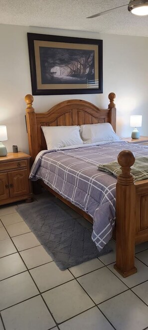 A bedroom with a wooden bed, a nightstand, a lamp, a framed picture on the wall, and a tiled floor.