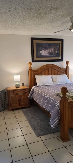 A bedroom with a wooden bed, a nightstand with a lamp, a framed picture on the wall, and a tiled floor.