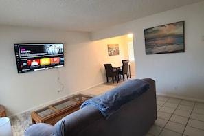 A living room with a flat-screen TV mounted on the wall, a grey sofa, a coffee table, and a dining area with chairs and a table.