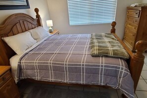 Una cama con cabecera, una mesita de noche con lámpara, un aparador y una ventana con persianas.