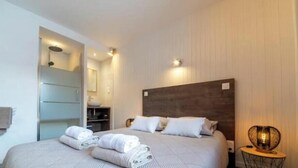 A hotel room with a bed, a bedside table, a lamp, and a bathroom visible through an open door. - Appt expo Sud 75 m2 - 1 séance SPA & Sauna - 3 Ch. (La Clusaz)
