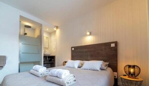 A hotel room with a bed, a bedside table, a lamp, and a bathroom visible through an open door. - Appt expo Sud 75 m2 - 1 séance SPA & Sauna - 3 Ch. (La Clusaz)