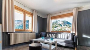 A room with a sofa, a table, and a view of a mountainous landscape through large windows. - Appt expo Sud 75 m2 - 1 séance SPA & Sauna - 3 Ch. (La Clusaz)