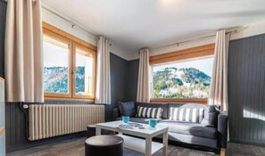 A room with a sofa, a table, and a view of a mountainous landscape through large windows. - Appt expo Sud 75 m2 - 1 séance SPA & Sauna - 3 Ch. (La Clusaz)