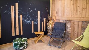 A room with a black wall, wooden slats, a green chair, a wooden stool, and a vase with branches. - Appt expo Sud 75 m2 - 1 séance SPA & Sauna - 3 Ch. (La Clusaz)