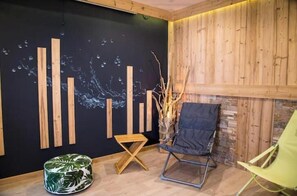A room with a black wall, wooden slats, a green chair, a wooden stool, and a vase with branches. - Appt expo Sud 75 m2 - 1 séance SPA & Sauna - 3 Ch. (La Clusaz)