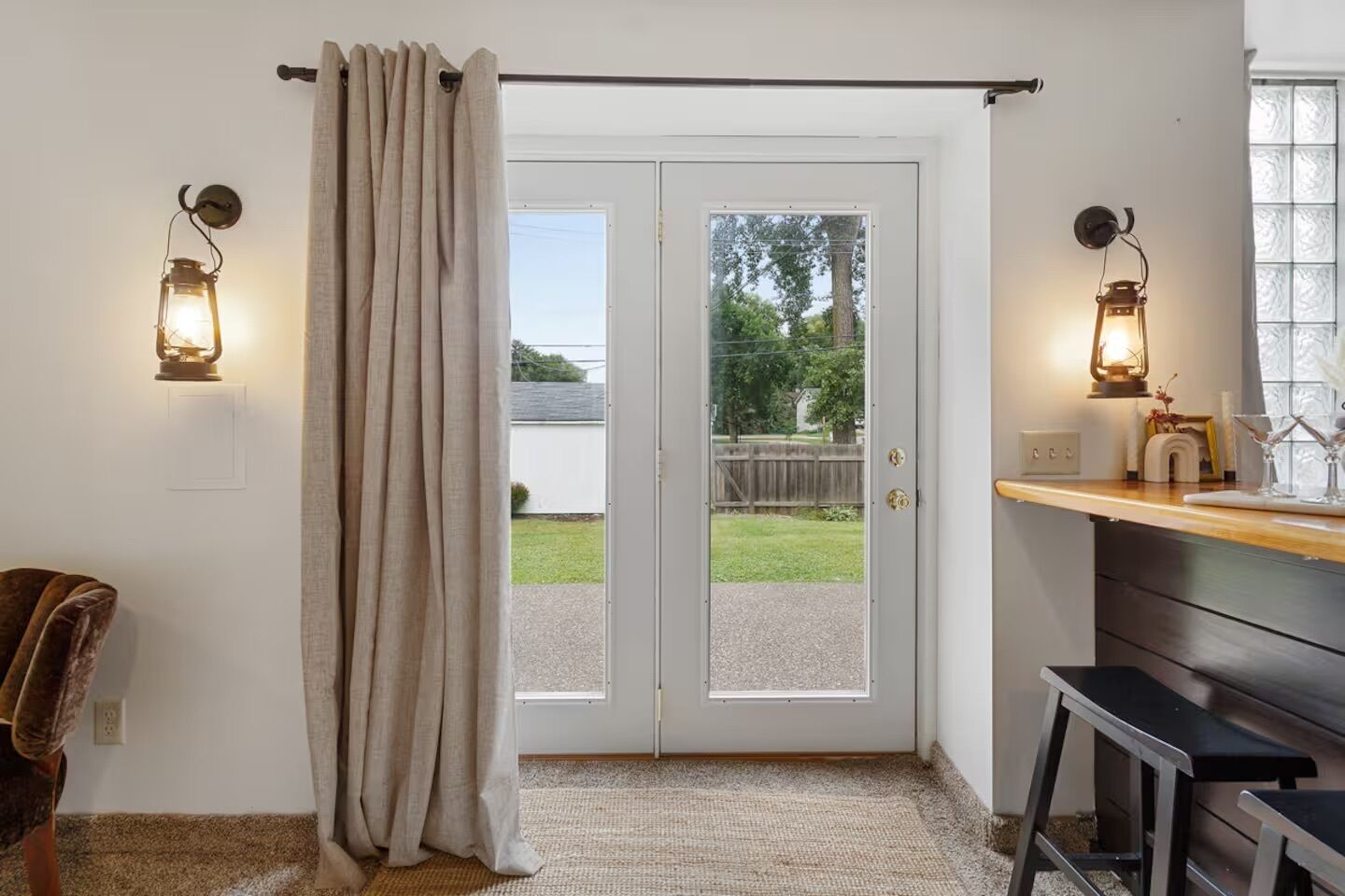 A room with a sliding door leading to a backyard, two wall-mounted lanterns, a wooden bench, and a small stool.