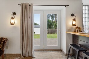 Ein Zimmer mit einer Schiebetür, die in einen Gartenwinkel führt, zwei Wandleuchten, eine Holzbank und ein kleiner Hocker.